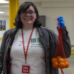 A person wearing glasses and a UNIQLO badge smiles while holding a mesh bag of small oranges, ready to help at the food bank. They are indoors in a white UNIQLO t-shirt, gray jacket, and one blue glove.