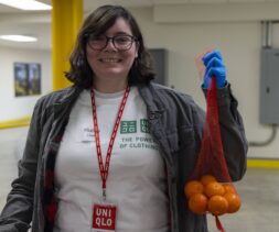 A person wearing glasses and a UNIQLO badge smiles while holding a mesh bag of small oranges, ready to help at the food bank. They are indoors in a white UNIQLO t-shirt, gray jacket, and one blue glove.