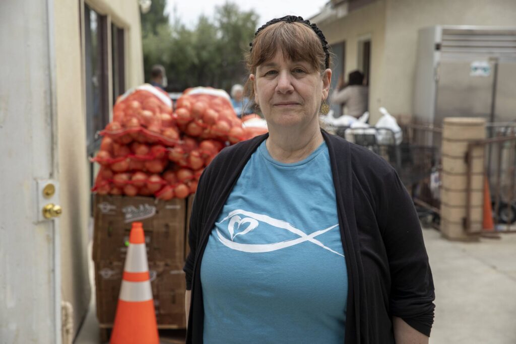 A woman wearing a blue shirt with a heart and fish logo stands outside near bags of onions stacked on pallets at a food bank, with a traffic cone and building in the background.