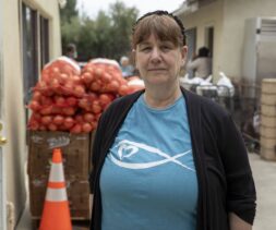 A woman wearing a blue shirt with a heart and fish logo stands outside near bags of onions stacked on pallets at a food bank, with a traffic cone and building in the background.