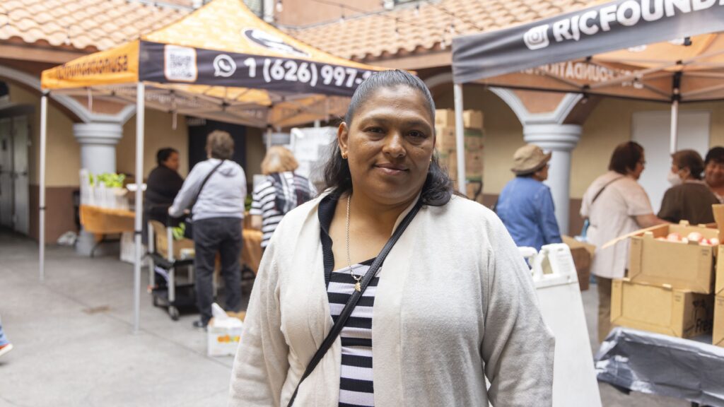 A woman in a light cardigan stands and smiles in front of outdoor market stalls, where people are gathered and boxes of produce—similar to those found at food pantries—are visible.