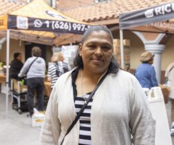 A woman in a light cardigan stands and smiles in front of outdoor market stalls, where people are gathered and boxes of produce—similar to those found at food pantries—are visible.