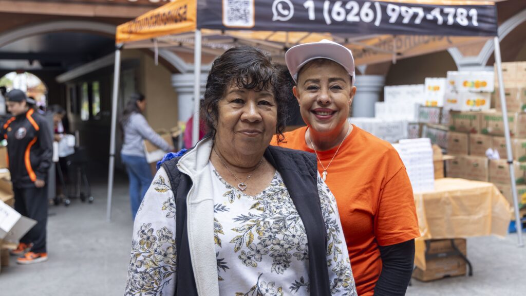 Two women smile at the camera at an outdoor food assistance event. One wears a floral-patterned top, the other an orange shirt and white cap. Tents, boxes, and other people are visible in the background.