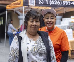 Two women smile at the camera at an outdoor food assistance event. One wears a floral-patterned top, the other an orange shirt and white cap. Tents, boxes, and other people are visible in the background.
