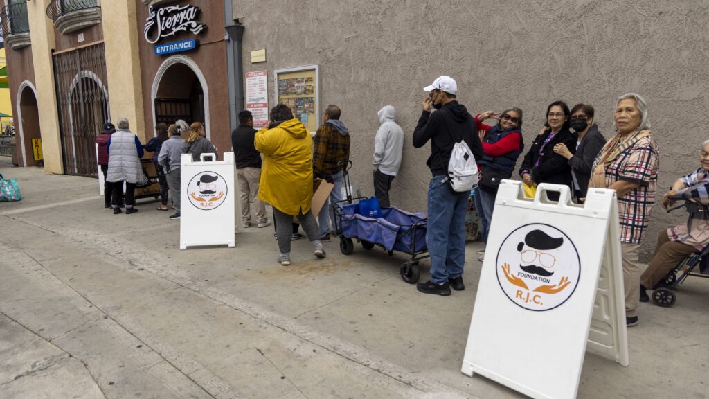 A diverse group of people stands in a long line on a sidewalk beside a building, possibly waiting for food assistance. Two sandwich boards with a logo featuring glasses and a mustache are visible. Some people are chatting or looking toward the camera.