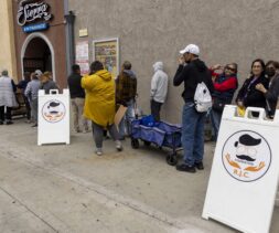 A diverse group of people stands in a long line on a sidewalk beside a building, possibly waiting for food assistance. Two sandwich boards with a logo featuring glasses and a mustache are visible. Some people are chatting or looking toward the camera.