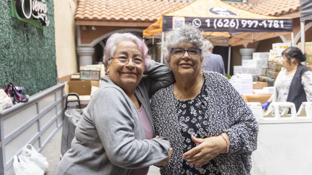 Two older women with gray hair, glasses, and casual clothing smile and pose together outdoors near a food pantry market stand. Other people and boxes are visible in the background.