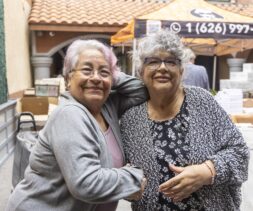 Two older women with gray hair, glasses, and casual clothing smile and pose together outdoors near a food pantry market stand. Other people and boxes are visible in the background.