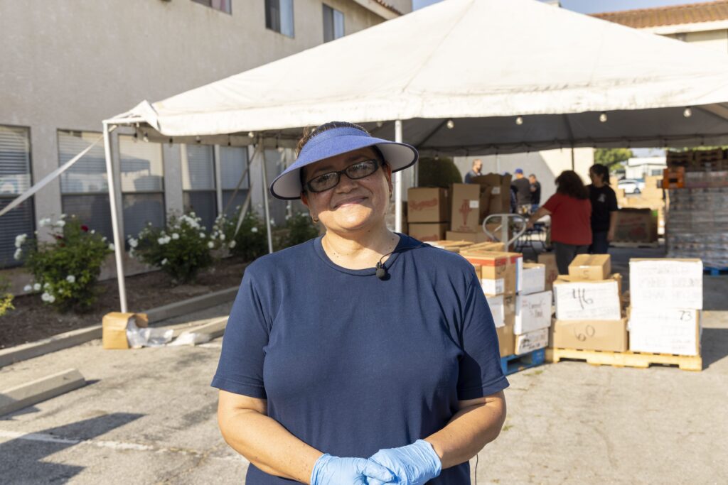 A woman wearing a blue shirt, gloves, and visor smiles while standing in front of a food bank tent with boxes and people organizing supplies outdoors.