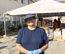 A woman wearing a blue shirt, gloves, and visor smiles while standing in front of a food bank tent with boxes and people organizing supplies outdoors.