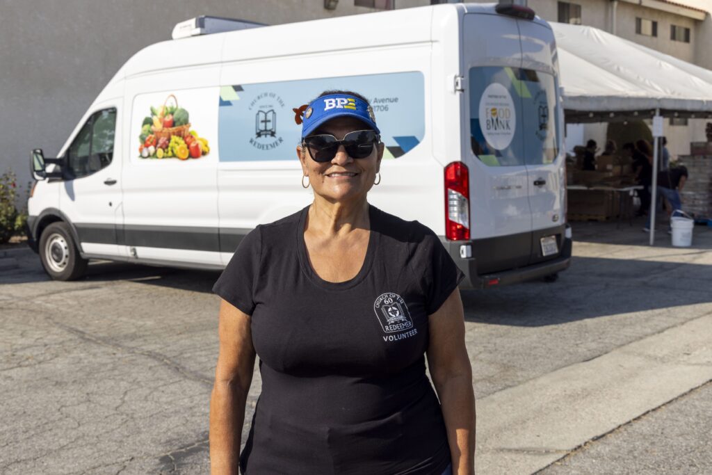 A woman wearing sunglasses, a blue cap, and a black volunteer shirt stands smiling in front of a food pantry van on a sunny day.
