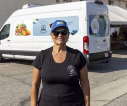 A woman wearing sunglasses, a blue cap, and a black volunteer shirt stands smiling in front of a food pantry van on a sunny day.