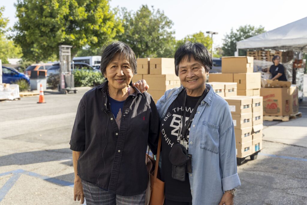 Two older women stand smiling outdoors in front of stacked cardboard boxes and a white tent at a food pantry. Trees and parked cars are visible in the background on a sunny day.