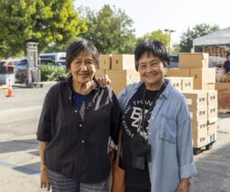 Two older women stand smiling outdoors in front of stacked cardboard boxes and a white tent at a food pantry. Trees and parked cars are visible in the background on a sunny day.