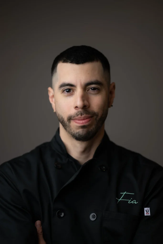 A man with short dark hair and a trimmed beard wears a black chef's coat embroidered with the name "Fia," standing against a plain, dark background.