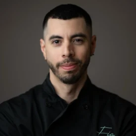 A man with short dark hair and a trimmed beard wears a black chef's coat embroidered with the name "Fia," standing against a plain, dark background.