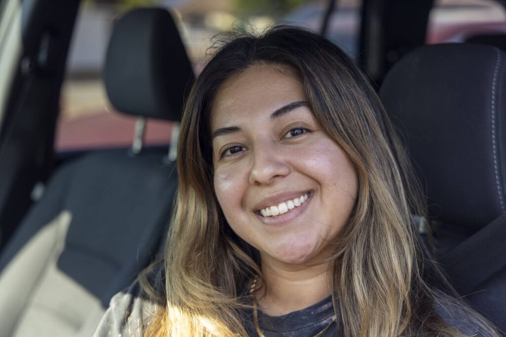 A woman with long brown hair smiles at the camera while sitting in a car, perhaps on her way to volunteer at a food pantry. She is wearing a dark shirt, and sunlight softly illuminates her face against the car’s visible interior.