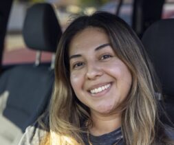 A woman with long brown hair smiles at the camera while sitting in a car, perhaps on her way to volunteer at a food pantry. She is wearing a dark shirt, and sunlight softly illuminates her face against the car’s visible interior.