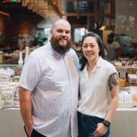 A smiling man and woman stand together in a modern shop or café, with shelves of snacks and products behind them. The woman has a visible tattoo on her arm and both wear light-colored shirts.