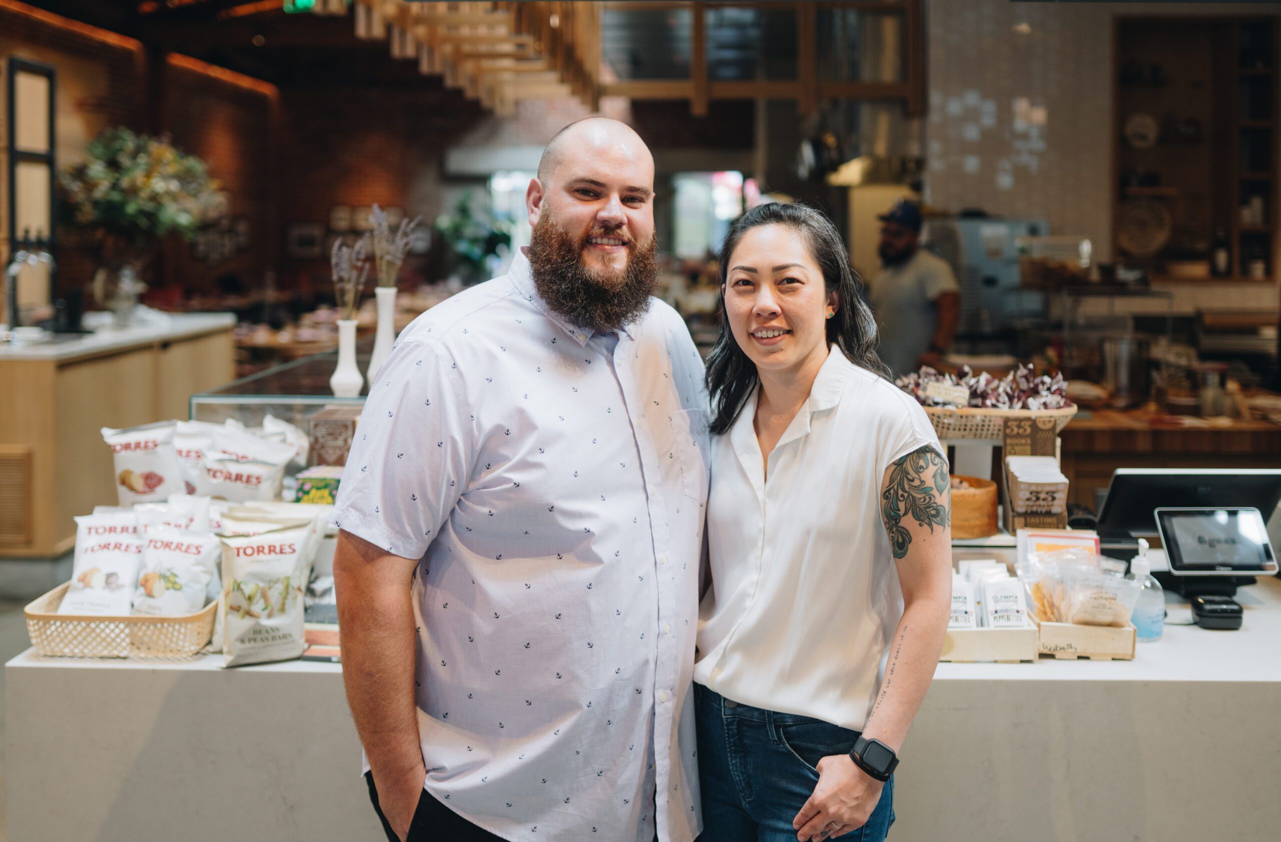 A smiling man and woman stand together in a modern shop or café, with shelves of snacks and products behind them. The woman has a visible tattoo on her arm and both wear light-colored shirts.