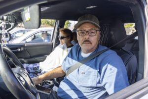 A man wearing glasses, a cap, and a blue shirt sits in the driver’s seat of a car, looking at the camera. Beside him, a woman with sunglasses sits in the passenger seat. They appear on their way to provide food assistance, both wearing seat belts.