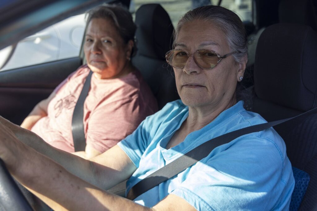Two older women sit in a car wearing seat belts, sunlight shining through the window. The driver, in glasses and a light blue shirt, and her friend in pink, discuss food assistance as they head to their next destination.
