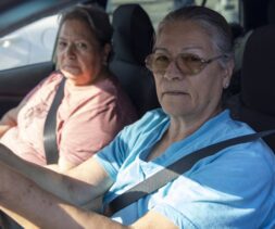 Two older women sit in a car wearing seat belts, sunlight shining through the window. The driver, in glasses and a light blue shirt, and her friend in pink, discuss food assistance as they head to their next destination.