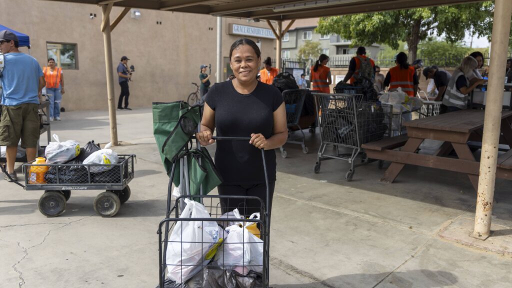 A woman smiles while standing outdoors with a cart full of bags, receiving food assistance. People in orange vests work in the background near shopping carts and tables under a canopy.