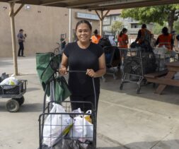 A woman smiles while standing outdoors with a cart full of bags, receiving food assistance. People in orange vests work in the background near shopping carts and tables under a canopy.