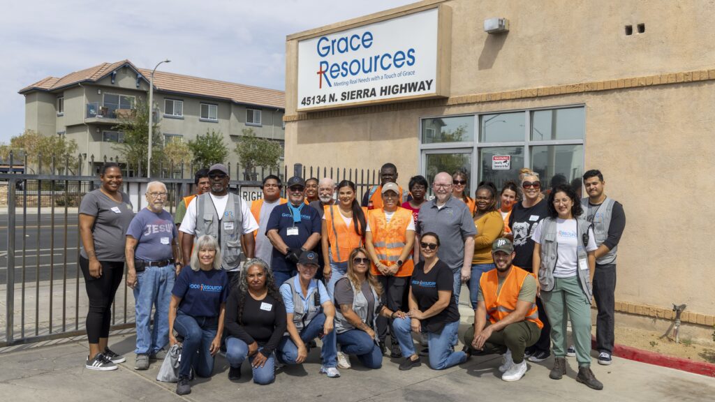 A diverse group of people, some wearing orange safety vests, stand and kneel together smiling outside a building with a 