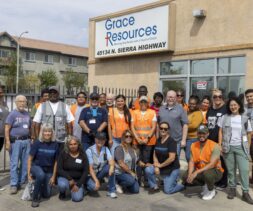 A diverse group of people, some wearing orange safety vests, stand and kneel together smiling outside a building with a 