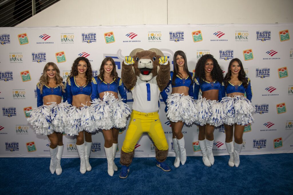 Seven cheerleaders in blue and white uniforms stand with a bear mascot in a football jersey and yellow pants, all smiling, holding white pom-poms, posing for a photo on the blue carpet at the Taste of the Rams event with branded backdrops behind them.