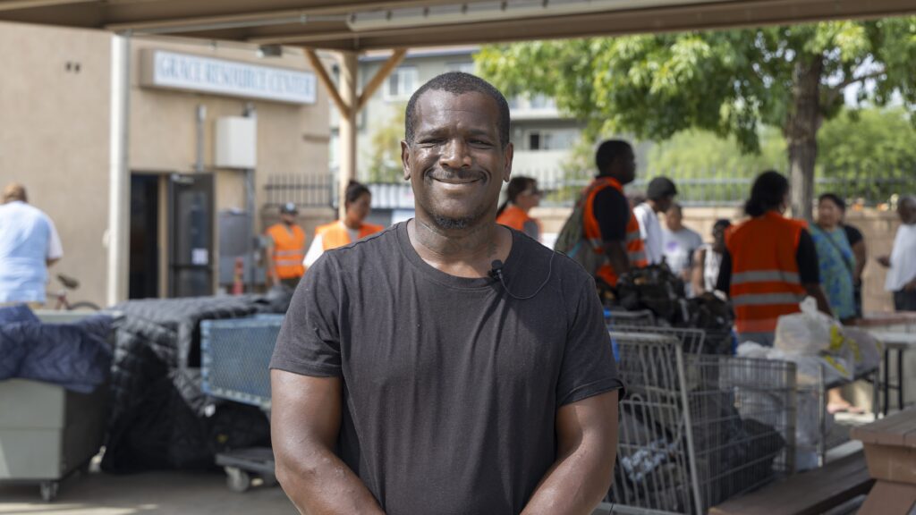 A man in a black t-shirt stands smiling outdoors, with a microphone clipped to his shirt. Behind him, several people in orange vests work near tables and carts at a food pantry under a shelter.