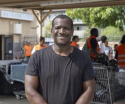 A man in a black t-shirt stands smiling outdoors, with a microphone clipped to his shirt. Behind him, several people in orange vests work near tables and carts at a food pantry under a shelter.