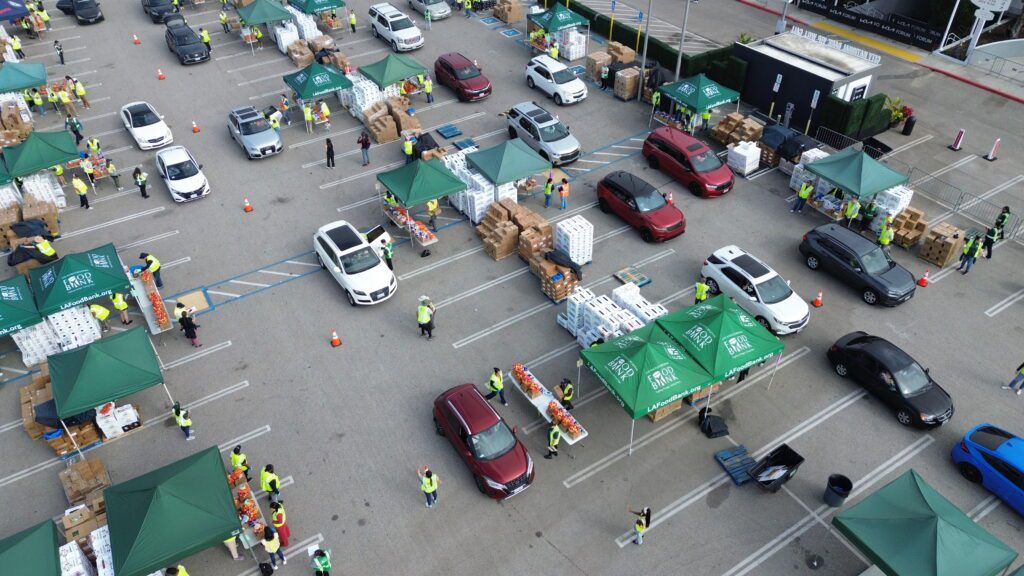 Aerial view of a large drive-through food assistance event in a parking lot, with cars lined up at green tented stations, volunteers in yellow vests, and stacks of boxes being handed out.