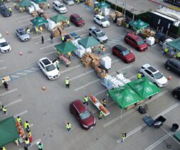 Aerial view of a large drive-through food assistance event in a parking lot, with cars lined up at green tented stations, volunteers in yellow vests, and stacks of boxes being handed out.
