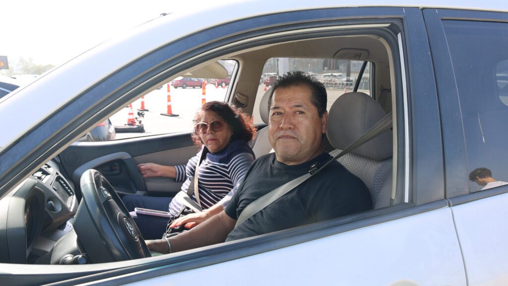 A man and a woman sit in the front seats of a white car, parked with the window down at a food assistance site. The man is in the driver's seat wearing a black shirt, while the woman wears sunglasses and a striped top. Traffic cones are visible outside.