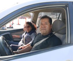 A man and a woman sit in the front seats of a white car, parked with the window down at a food assistance site. The man is in the driver's seat wearing a black shirt, while the woman wears sunglasses and a striped top. Traffic cones are visible outside.