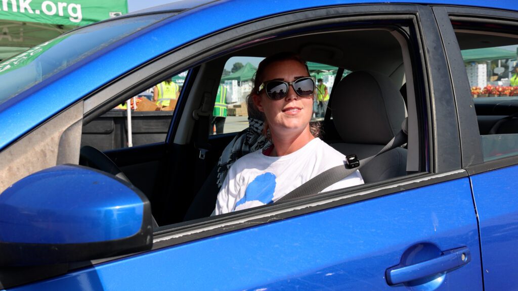 A person wearing sunglasses and a white t-shirt sits in the driver’s seat of a blue car with the window down on a sunny day, ready to volunteer at the local food bank.
