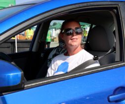 A person wearing sunglasses and a white t-shirt sits in the driver’s seat of a blue car with the window down on a sunny day, ready to volunteer at the local food bank.