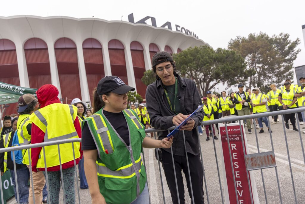 Two event staff in safety vests stand by a barricade, one pointing at a clipboard. Behind them, a crowd in yellow vests—likely food bank volunteers—gathers outside the Kia Forum arena.