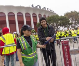 Two event staff in safety vests stand by a barricade, one pointing at a clipboard. Behind them, a crowd in yellow vests—likely food bank volunteers—gathers outside the Kia Forum arena.