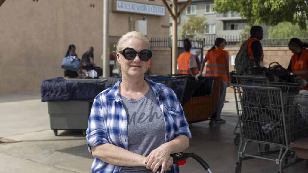 A woman with blonde hair in a ponytail, wearing large black sunglasses, a plaid shirt, and a gray t-shirt, stands outdoors near a food pantry, with people and shopping carts visible in the background by the resource center.