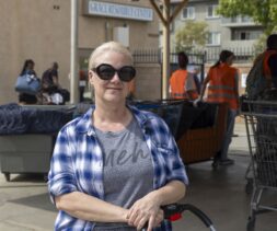 A woman with blonde hair in a ponytail, wearing large black sunglasses, a plaid shirt, and a gray t-shirt, stands outdoors near a food pantry, with people and shopping carts visible in the background by the resource center.