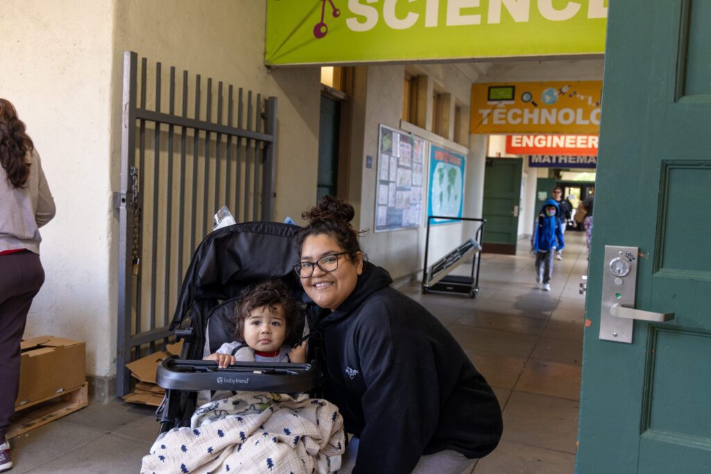 A woman crouches next to a stroller with a young child under a blanket in a school hallway. A green sign overhead reads 