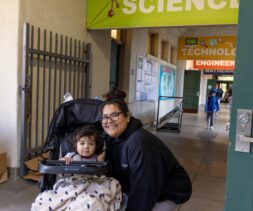 A woman crouches next to a stroller with a young child under a blanket in a school hallway. A green sign overhead reads 