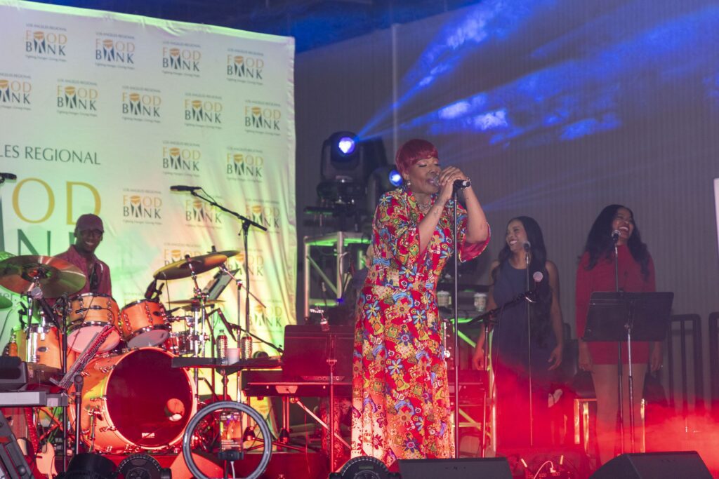 A woman in a colorful floral outfit sings on stage with a live band, joined by two backup singers. Bright stage lights and Food Bank banners are in the background at this Los Angeles Clippers community event.