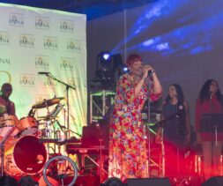 A woman in a colorful floral outfit sings on stage with a live band, joined by two backup singers. Bright stage lights and Food Bank banners are in the background at this Los Angeles Clippers community event.