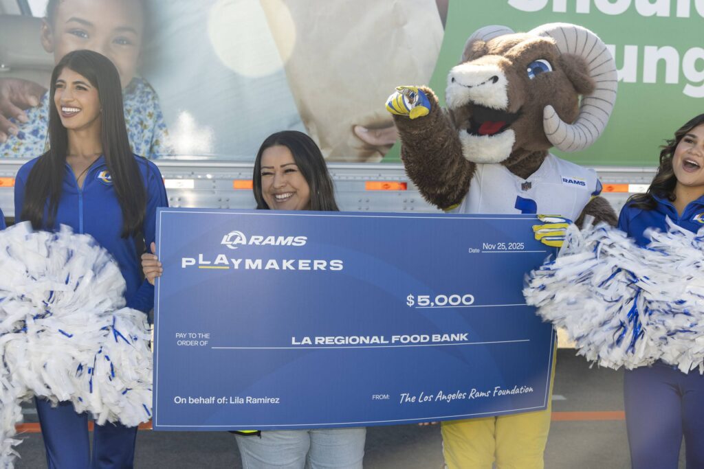 Three smiling women and the Los Angeles Rams mascot hold a large $5,000 check from the Rams Playmakers to LA Regional Food Bank. Two women hold pompoms, celebrating their support for the food bank, with a branded truck in the background.