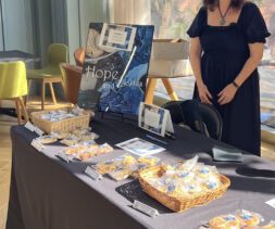 A woman in a black dress stands behind a table covered with baskets of cookies for sale. A sign reads 